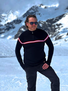  Man wearing a black sweater with red and white stripes and sunglasses, standing in a snowy mountain landscape.