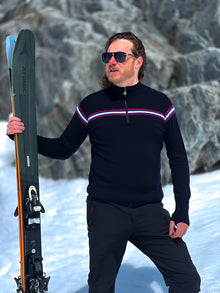  Man wearing navy ski jumper with a chest stripe in snowy background holding skis